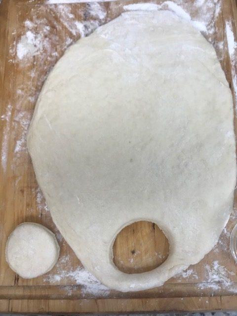 A sheet of dough, ready for parker house rolls, is rolled out on a floured wooden surface, with a round piece cut out and placed nearby and another circular cut-out in the main dough sheet.