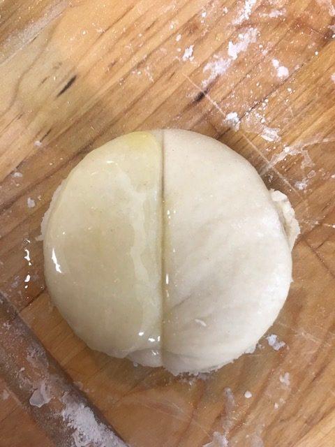 A round ball of dough on a floured wooden surface, with one half covered in a shiny layer of oil and the other half plain, ready to be shaped into classic Parker House rolls.