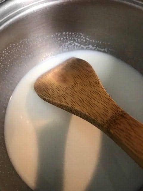 A close-up of a wooden spatula stirring milk in a stainless steel pot, preparing the base for fluffy parker house rolls.