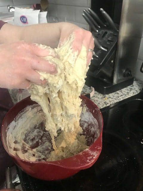 A person’s hands stretch and knead sticky Parker House rolls dough above a red mixing bowl in a kitchen, with flour and kitchen utensils visible in the background.