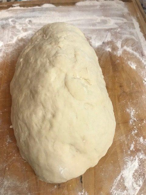 A ball of raw bread dough sits on a floured wooden surface, ready to be shaped into soft, buttery Parker House rolls or baked to perfection.