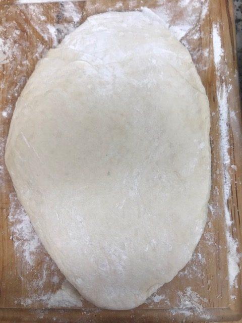 A piece of dough, dusted with flour, is rolled out into an oval shape on a wooden surface sprinkled with flour—reminiscent of how parker house rolls are prepared before baking.