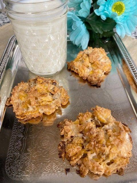 Three homemade Corn Flake Macaroons sit on a silver tray next to a glass jar of milk. Blue flowers in the background add a touch of color to the cozy scene.