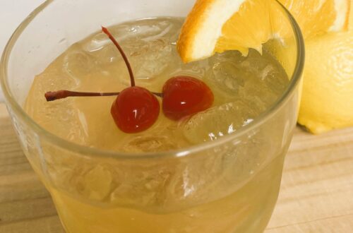 Whiskey Sour in clear glass with 2 maraschino cherries and orange slice.