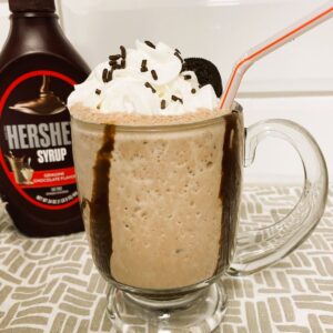 Chocolate Milkshake with whipped cream and sprinkles in clear glass with chocolate syrup bottle in background.