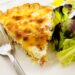 Slice of quiche on white plate next to fork and green salad.