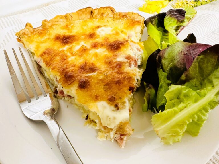 A slice of quiche with a golden, cheesy top is served on a white plate next to a small side salad of mixed greens, offering inspiration for anyone interested in how to make Quiche Lorraine. A fork rests neatly on the plate.