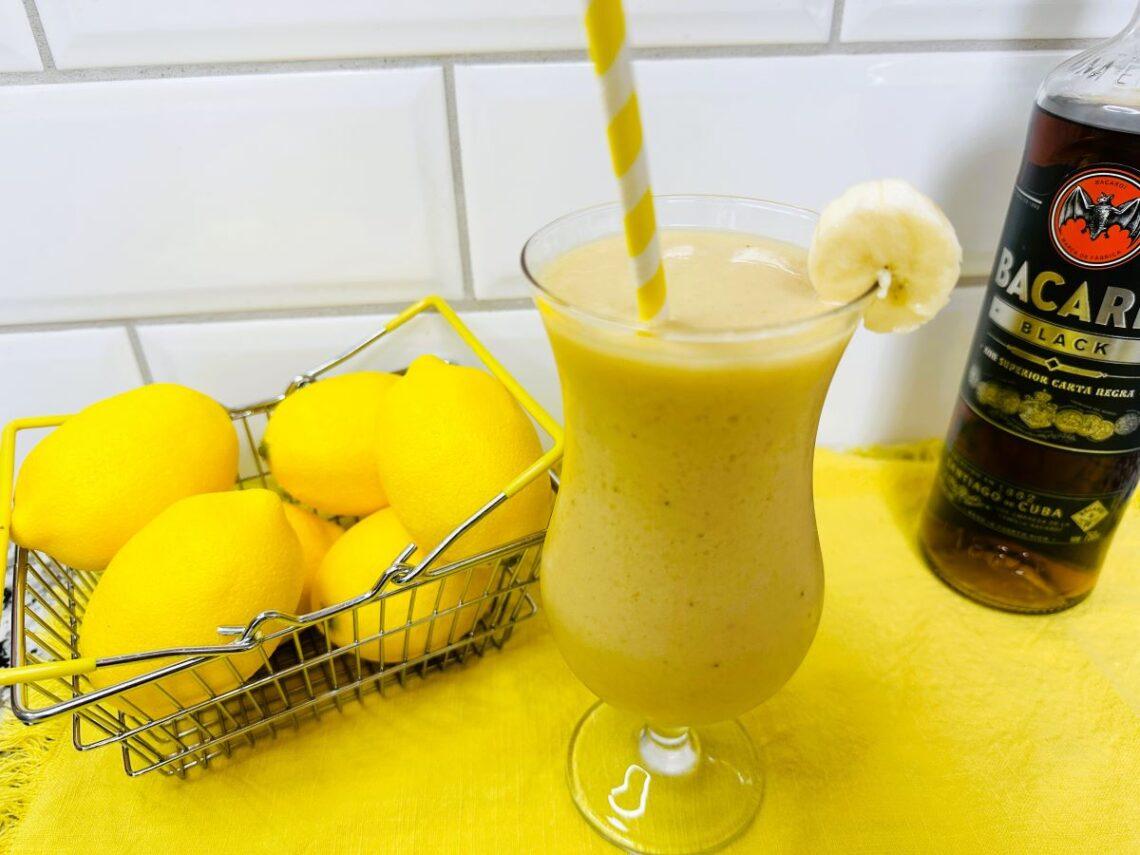 Banana daiquiri in hurricane glass with lemons and rum in background.