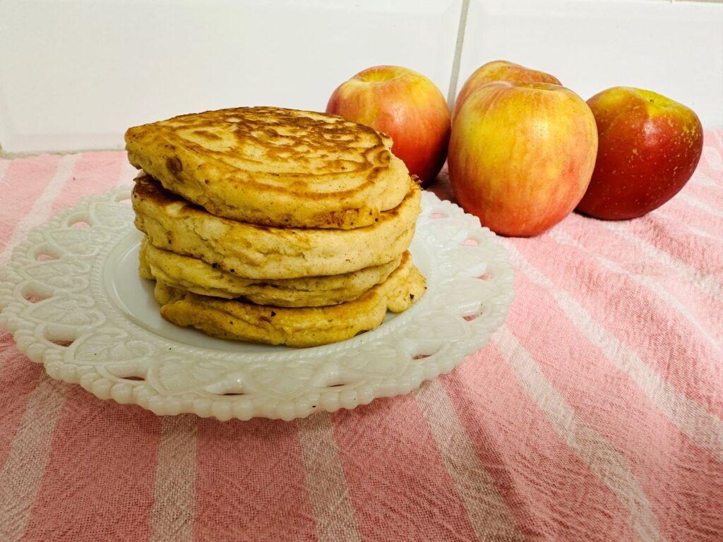 Stack of apple pancakes on white plate with four apples in background.