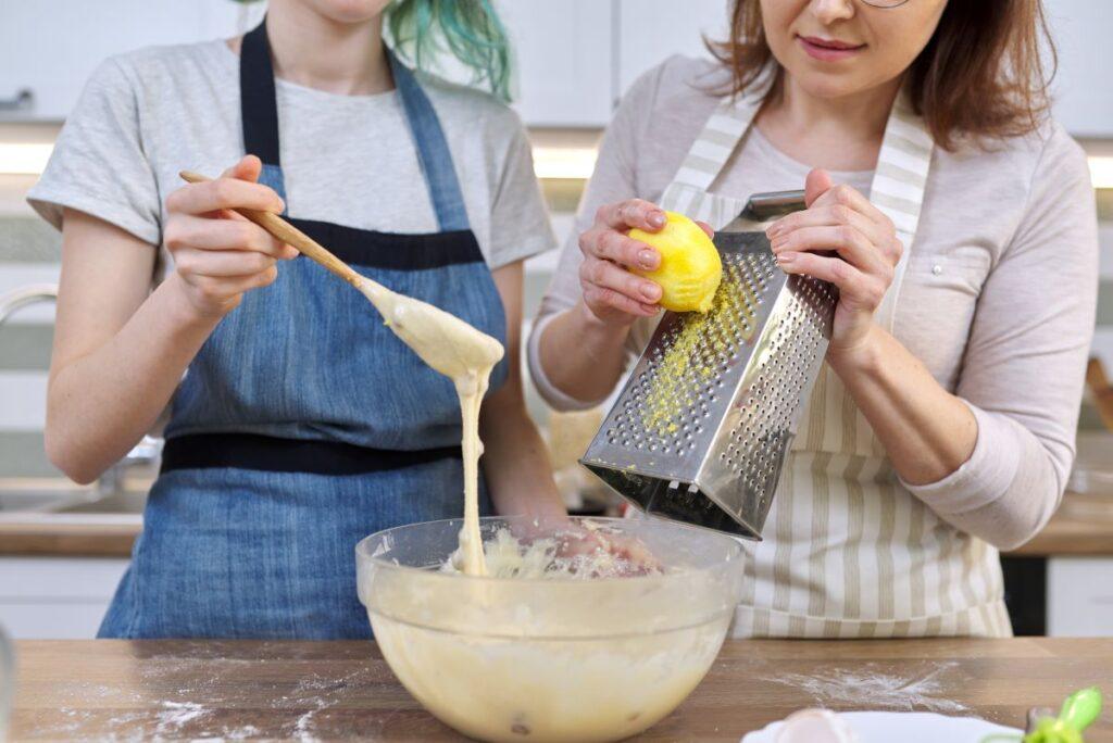 Two women zesting a lemon with a box grater into batter.