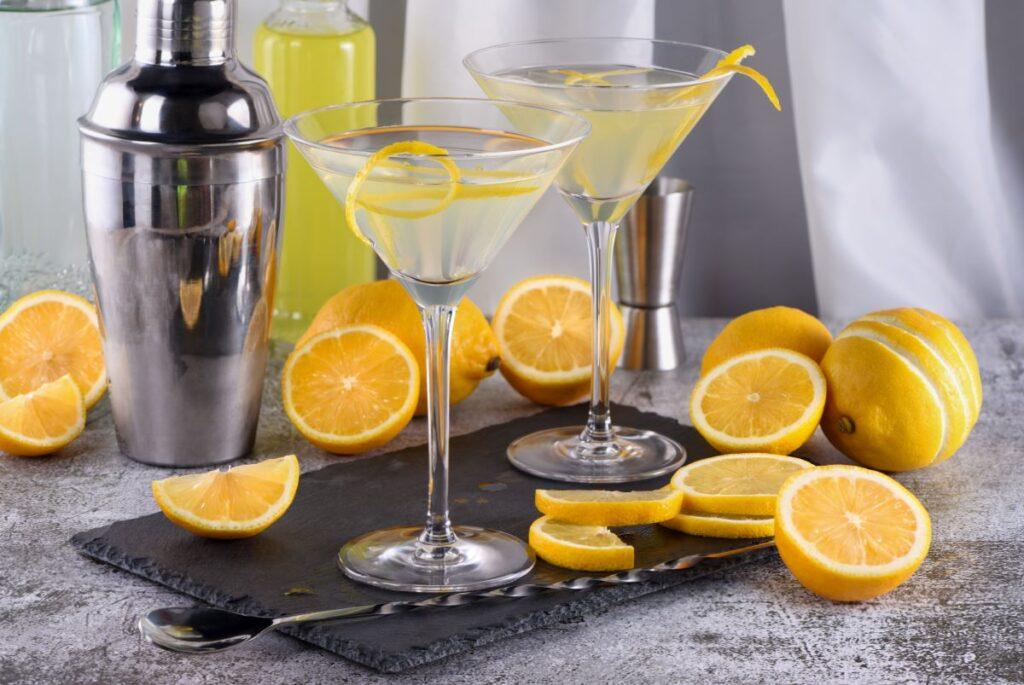 Lemon drop martinis containing lemon zest with sliced lemons and martini shaker in background.