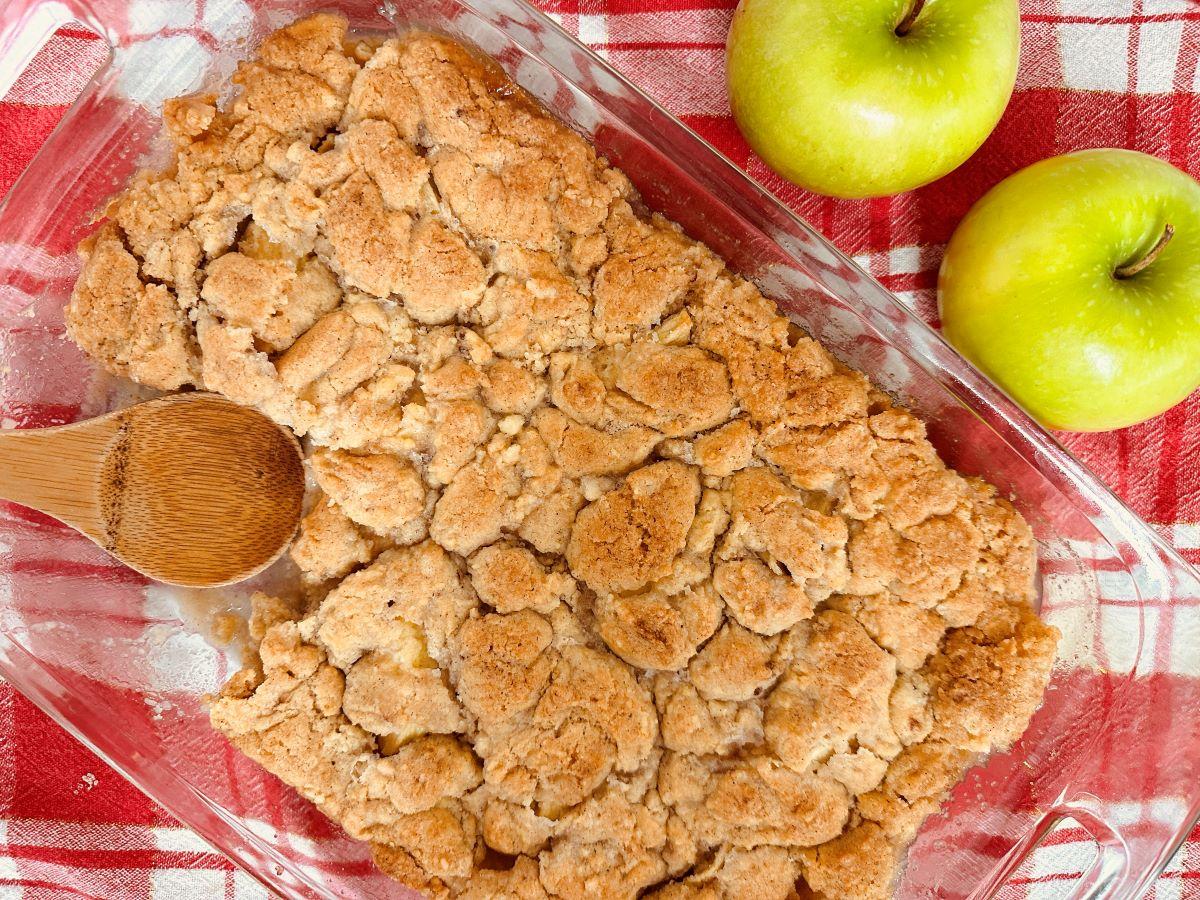 Apple crisp in clear dish with one serving missing and green apples in background.