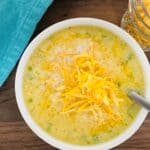 A bowl of creamy cheeseburger chowder topped with shredded cheddar cheese, a spoon inside. A blue cloth sits beside the bowl and a small glass jar filled with more shredded cheese is nearby, all on a wooden surface.