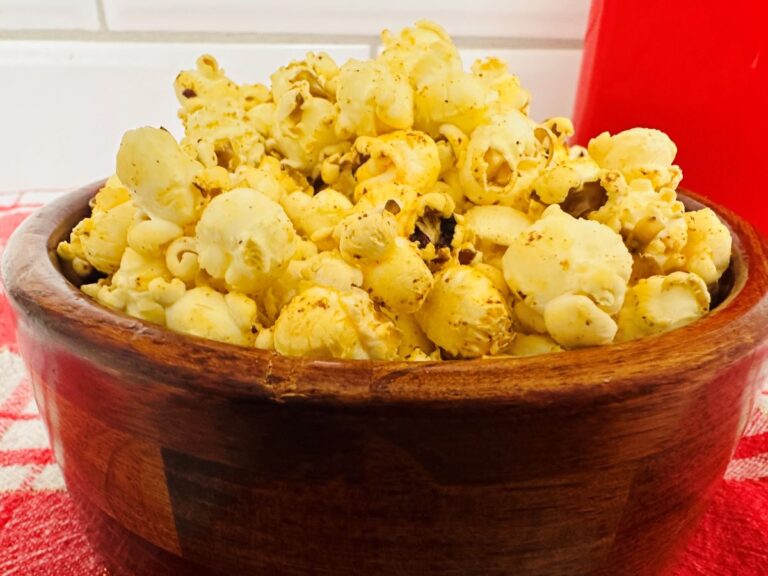 A wooden bowl filled with Chili Popcorn sits on a red and white checkered cloth, with a red container and white tiled wall in the background.