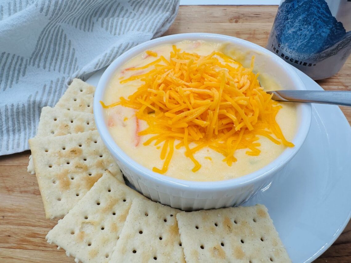 Yellow soup with shredded cheddar cheese on top in white bowl. Soda crackers on plate underneath.