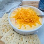 A white bowl filled with creamy cheddar cheese beer soup, topped with shredded cheddar, sits on a plate surrounded by saltine crackers. A spoon is in the bowl, and a striped towel and a drink can are in the background.