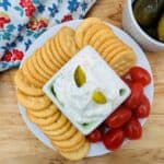 A plate with round crackers, cherry tomatoes, and a bowl of creamy Dill Pickle Dip topped with pickle slices. A bowl of pickles and a colorful floral napkin are nearby on a wooden surface.