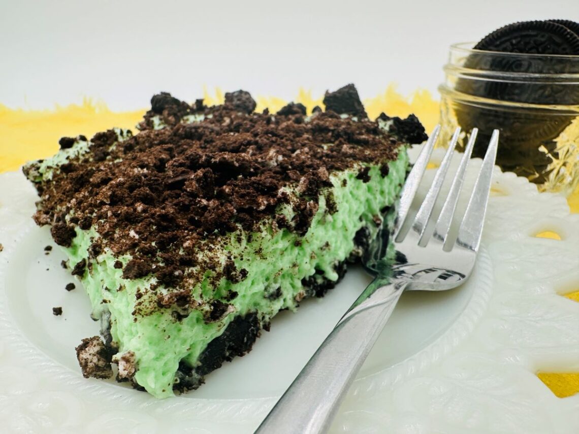 Green slice of pie with dark brown Oreo crumbs on top on white plate.