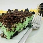 A slice of mint Oreo dessert, reminiscent of grasshopper pie with its green creamy filling and crushed Oreo topping, sits on a white plate with a fork. In the background, a small jar filled with Oreos rests on a yellow surface.