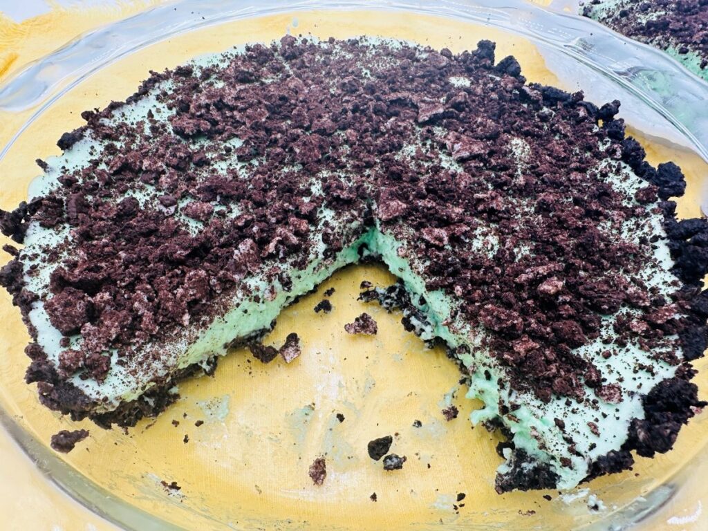 Green pie with Oreo crumbs on top with one slice missing in glass pie dish.