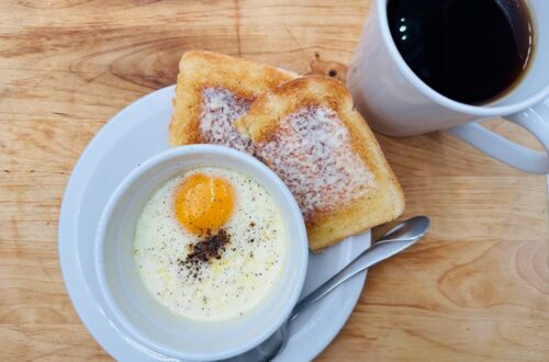 Cooked egg in white cup on top of white plate with buttered toast and white mug of coffee in background.