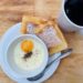 Cooked egg in white cup on top of white plate with buttered toast and white mug of coffee in background.
