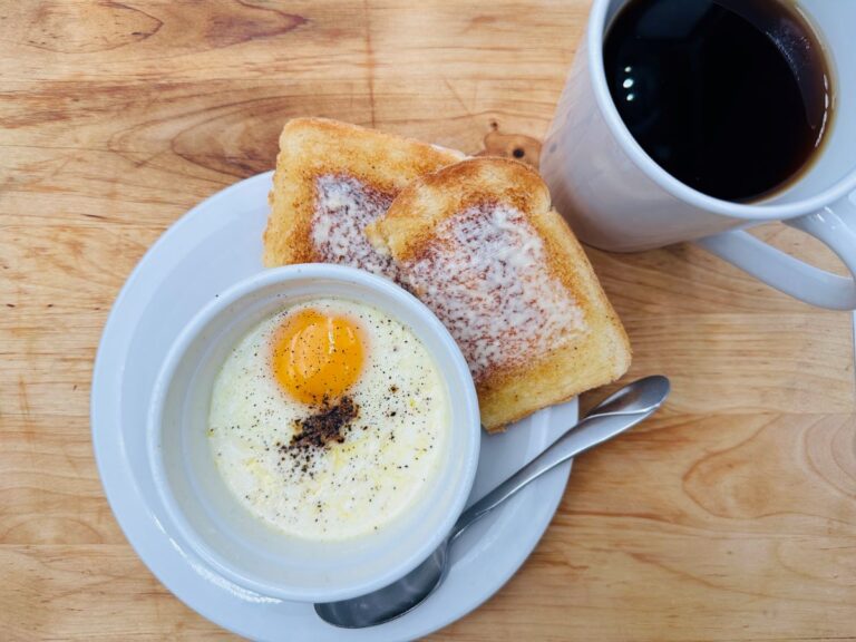 Cooked egg in white cup on top of white plate with buttered toast and white mug of coffee in background.