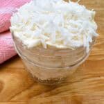 White frosting with shredded coconut on top in clear glass jar.
