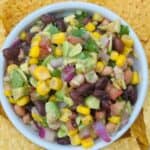 Bean, corn, and onion mixture in white bowl surrounded by tortilla chips.