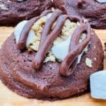Close up of chocolate cookies with mini marshmallows, chopped walnuts, and drizzled chocolate frosting on wooden background with mini marshmallows scattered around.