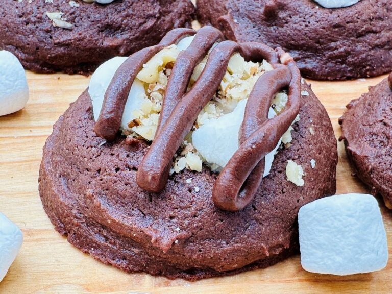 Close up of chocolate cookies with mini marshmallows, chopped walnuts, and drizzled chocolate frosting on wooden background with mini marshmallows scattered around.