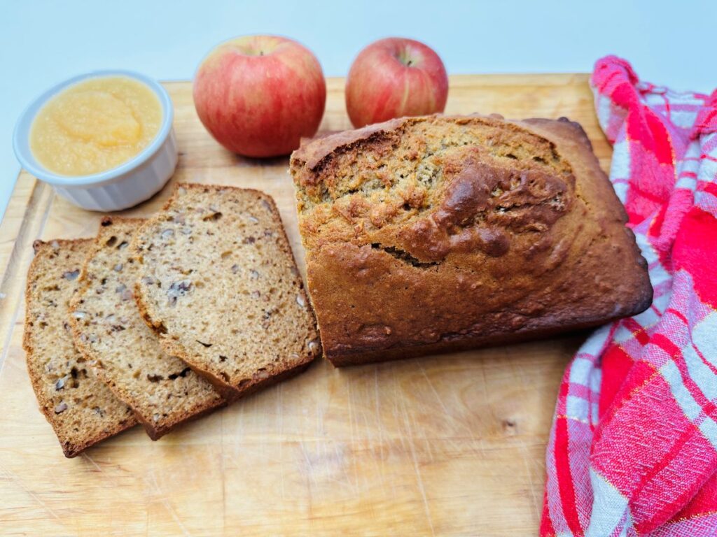 Three slices of bread next to the rest of the loaf on wooden board with white bowl of applesauce and two red apples in the background.
