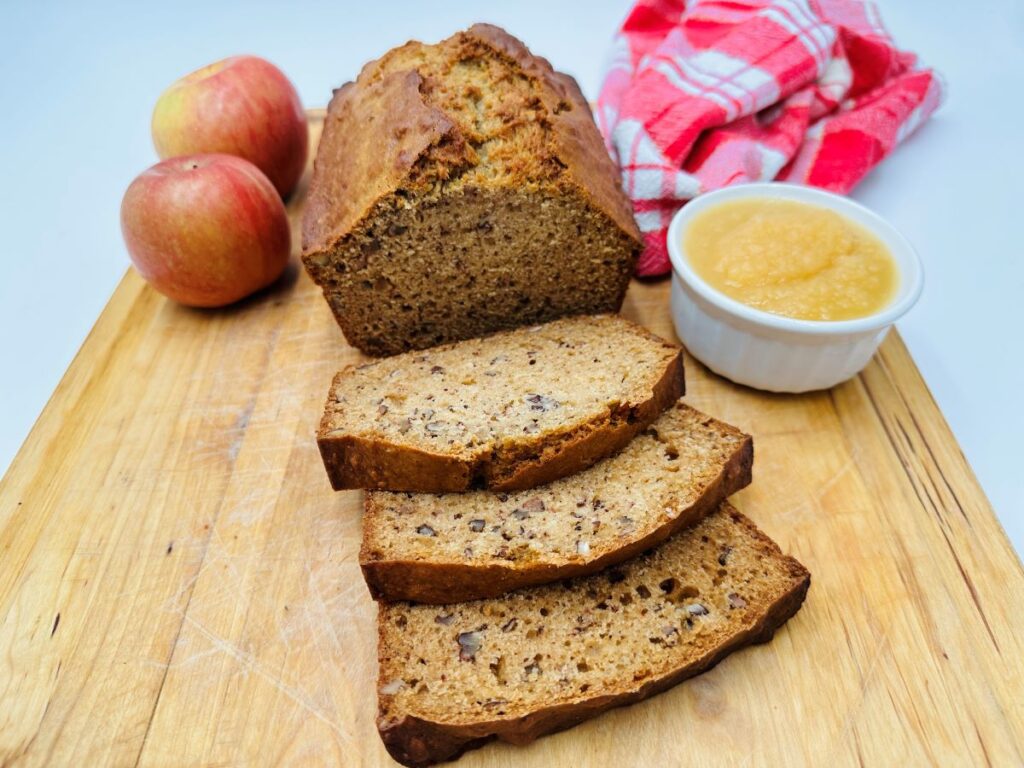 Three slices of bread next to the rest of the loaf on wooden board with white bowl of applesauce and two red apples in the background.
