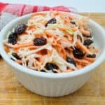 Mixture of shredded carrots, shredded cabbage, and raisins topped with white dressing in white bowl on wooden surface with red and white checkerd napkin in the background.