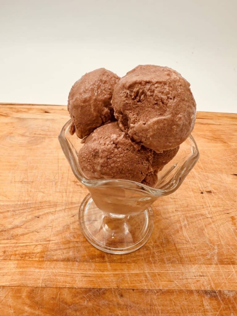 Three scoops of chocolate ice cream in glass sundae dish on wooden board.