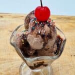 Three scoops of chocolate ice cream with colorful sprinkles and a cherry on top in glass sundae dish.