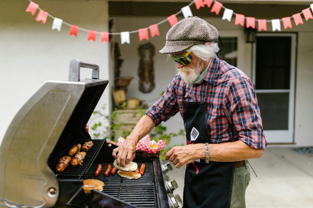 Man grilling hot dogs and hot dogs bun on outside grill.