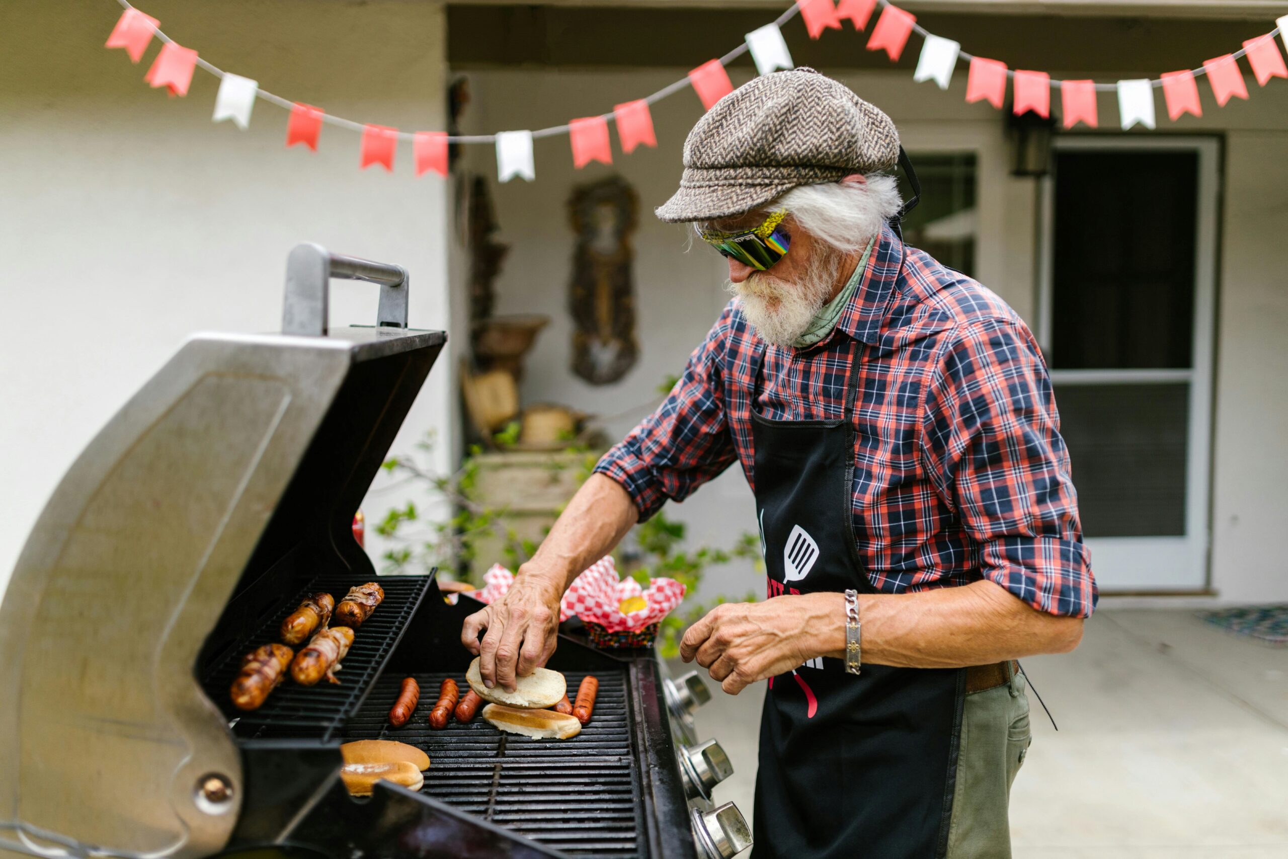 Man grilling hot dogs and hot dogs bun on outside grill.