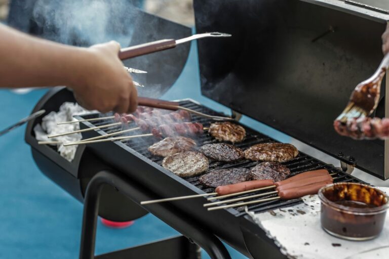 Hand holding tongs over barbecue grill full of hamburgers and hot dogs.