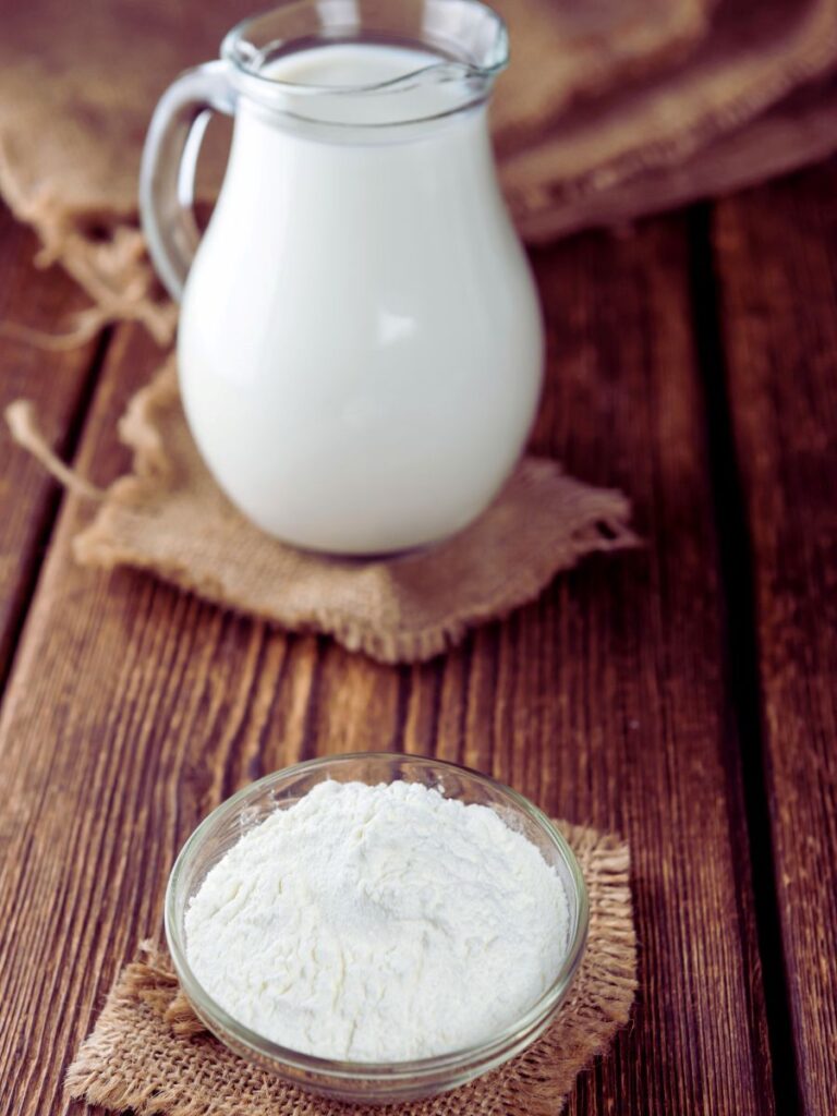 White powder in clear dish in front of glass pitcher of milk.