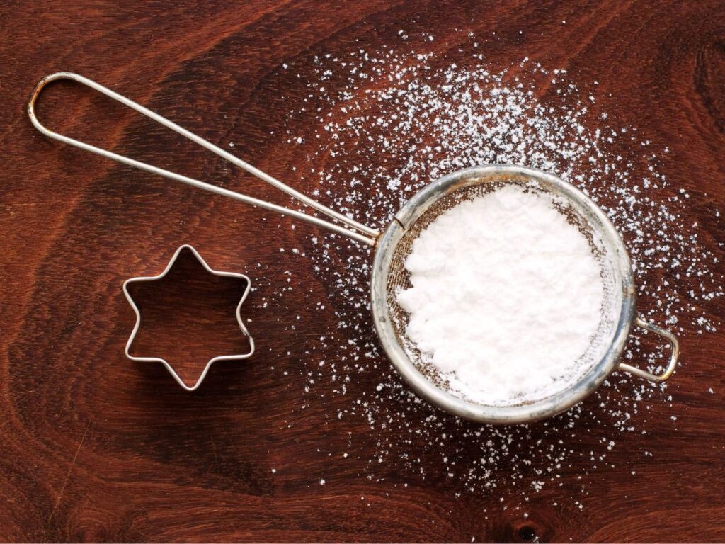 Powdered sugar in silver circle next to star cookie cutter on wooden surface.