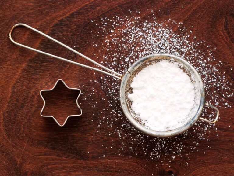 Powdered sugar in silver circle next to star cookie cutter on wooden surface.