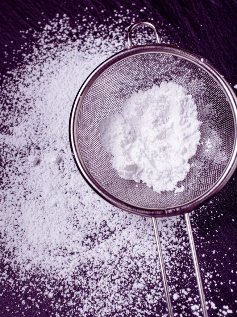 Powdered sugar in silver strainer above black surface.