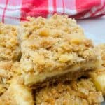Close up of cheesecake bar with light brown crust, white filling, and crumb topping on top of a layer of more cheesecake bars.