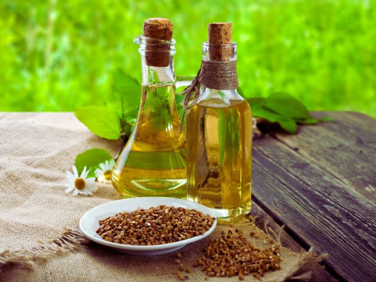 Two glass bottles of yellow oil with corks on top behind white dish of seeds.