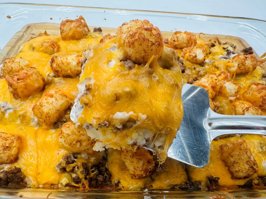 One serving of cheesy tater tot casserole on silver serving spatula held above cheesy casserole.