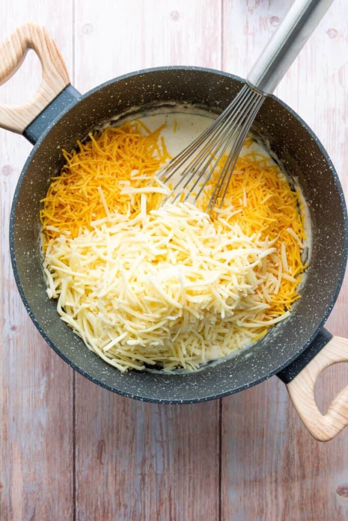 White and yellow grated cheese in white sauce in gray pot with whisk.