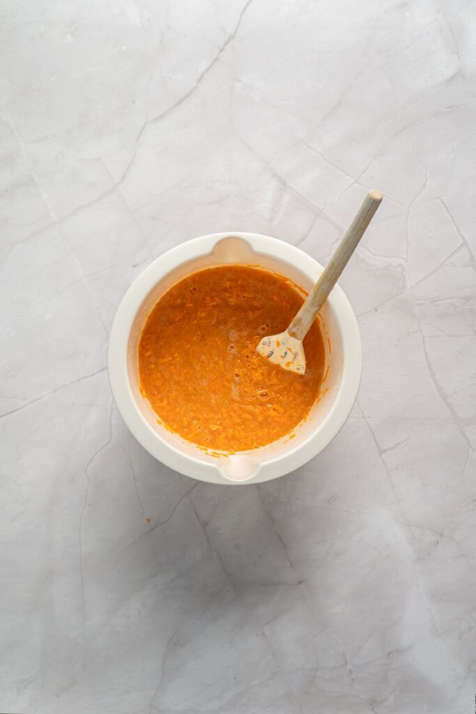 Orange mixture with small carrot shreds with rubber spatula in white bowl.