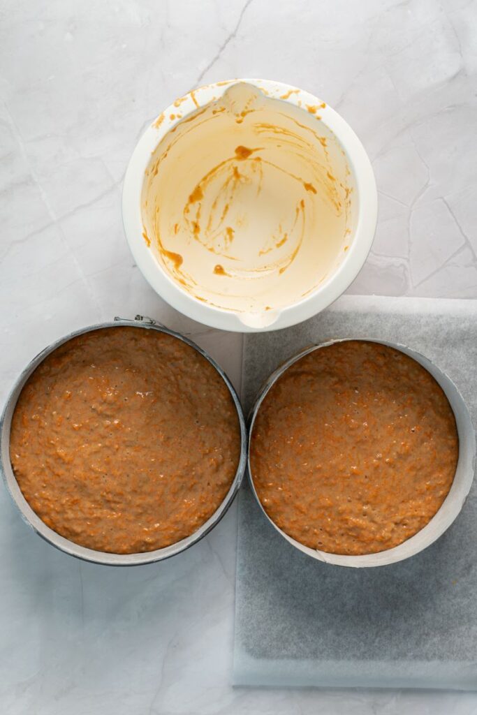 Top view of empty white bowl with two pans of light brown cake batter in silver cake pans below.