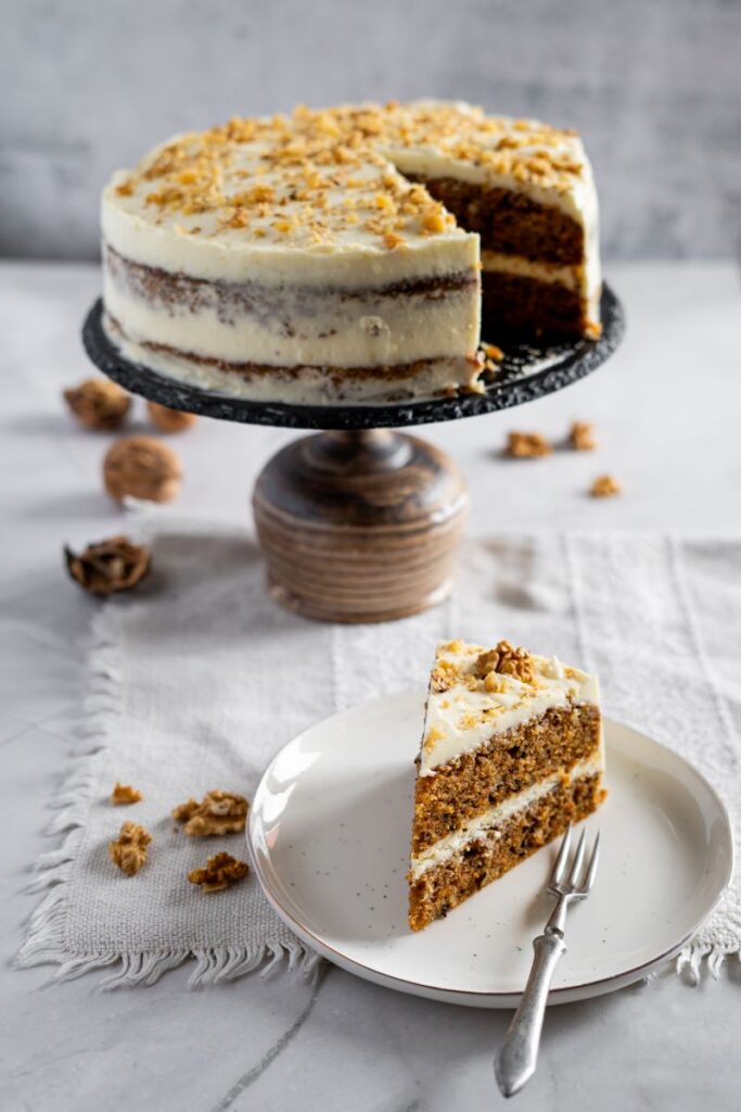 Slice of carrot cake on white plate in front of whole carrot cake with one slice missing.